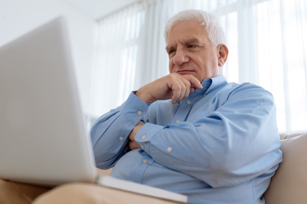 Elderly man sitting on comfy sofa and researching furnace noise on laptop.