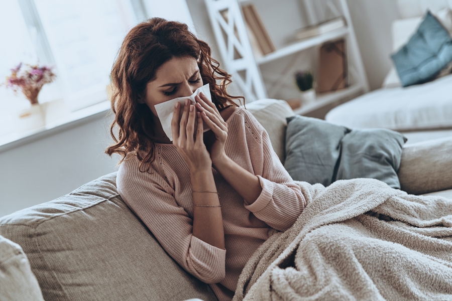 Top view of sick young women blowing her nose using facial tissues while sitting on the sofa at home.